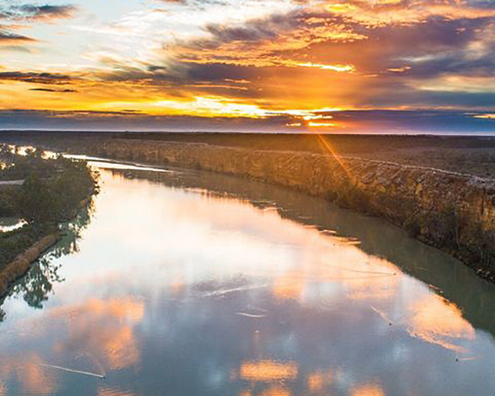 The Murray river curves into the distance, reflecting a bright, orange sunset in a drone photograph. Photo credit: Ben Stamatovich
